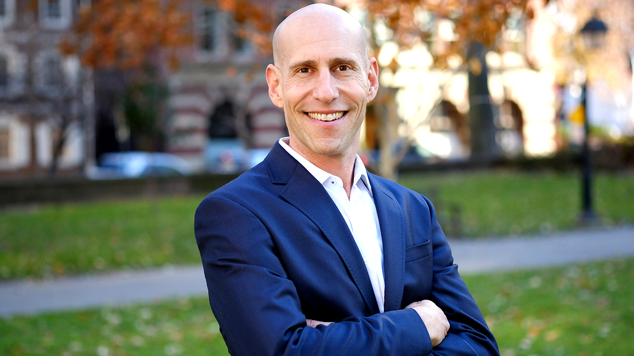 Headshot of Brian Shapiro wearing a dark blue blazer and white button-up shirt as he stands in a park in the fall with gold leaves scattered on the ground